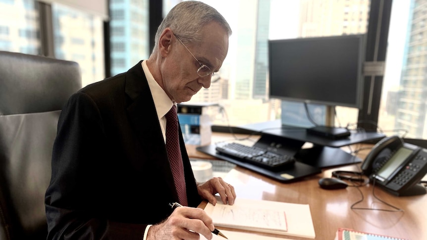 ACCC 主席 Rod Sims 坐在在他的办公桌上用笔标记文件。